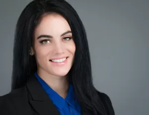 Professional Photographer A woman with long black hair wearing a black blazer and blue blouse smiles at the camera against a plain gray background, capturing a classic style often seen in professional portraits. www.maranonimagery.com