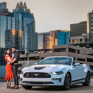 Professional Photographer A couple stands close together on a rooftop parking lot, with a silver Ford Mustang convertible in front and tall modern buildings behind them at sunset—captured in stunning wedding photography by a professional photographer. www.maranonimagery.com