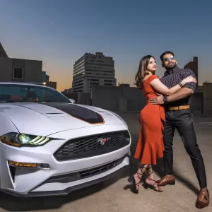 Professional Photographer A stylish couple poses on a rooftop parking lot at sunset beside a white Ford Mustang. Perfect for a photographer seeking dynamic city scenes, the woman stuns in orange as the man sports sunglasses and a patterned shirt with buildings behind them. www.maranonimagery.com