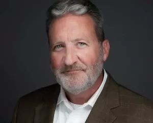 Professional Photographer A middle-aged man with gray hair and beard, wearing a brown suit jacket and white shirt, smiles slightly while posing for professional headshots against a dark gray background. www.maranonimagery.com