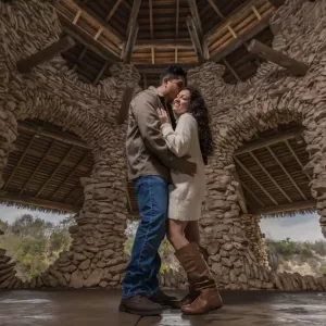 Professional Photographer A couple embraces and smiles inside a rustic stone pavilion with wooden beams and arches, standing close together on a polished floor—captured beautifully through the artistry of wedding photography. www.maranonimagery.com