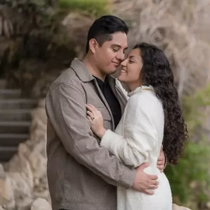 Professional Photographer A couple stands close together outdoors, hugging and smiling with their eyes closed. Perfect for portraits or headshots, the scene features a brown jacket, white sweater, stone steps, and greenery in the background. www.maranonimagery.com