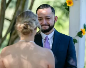 Professional Photographer A man in a suit with a purple tie smiles emotionally at a woman in a white dress during an outdoor wedding ceremony, with sunflowers and greenery in the background, beautifully captured by a skilled wedding photographer. www.maranonimagery.com