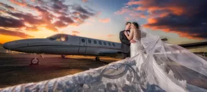 Professional Photographer A bride and groom embrace in front of a private jet at sunset as their wedding photographer captures the scene, with the bride’s long lace veil flowing dramatically toward the camera and colorful clouds filling the sky. www.maranonimagery.com