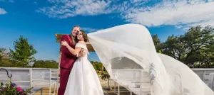 Professional Photographer A bride and groom embrace outdoors under a blue sky with wispy clouds, captured by a skilled photographer. The bride’s long veil flows dramatically as benches and greenery surround them, both smiling joyfully in this perfect headshot moment. www.maranonimagery.com