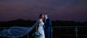 Professional Photographer A bride and groom stand close together on a balcony at dusk. The bride’s veil flows in the wind, and she holds a bouquet of blue flowers. This romantic wedding photography scene is set against a deep blue and purple sky with trees in the distance. www.maranonimagery.com