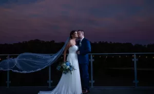 Professional Photographer A bride and groom stand on a balcony at dusk, captured in elegant wedding photography. The bride’s white dress and blue bouquet complement the groom’s navy suit against a sky of purple and pink hues. www.maranonimagery.com