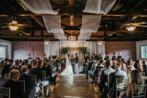 Professional Photographer A wedding ceremony inside a decorated hall with draped fabric and chandeliers. The photographer captures stunning wedding photography as the bride and groom stand at the altar with their party, while guests sit on gold chairs facing them. www.maranonimagery.com