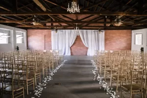 Professional Photographer Rows of gold chairs face a simple wedding altar with white drapes in a rustic hall with exposed wooden beams and brick walls—an inviting setting perfect for a photographer to capture unforgettable moments amid chandeliers and flower petals lining the aisle. www.maranonimagery.com