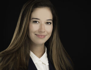 Professional Photographer A young woman with long brown hair smiles softly at the camera in this professional headshot. She is wearing a white collared shirt and a dark blazer, posed against a solid black background. www.maranonimagery.com