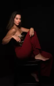 Professional Photographer A woman with long hair sits sideways on a black chair against a dark background, dressed in a strapless top and burgundy pants. Her elegant, confident expression is perfect for professional headshots captured by an experienced photographer. www.maranonimagery.com
