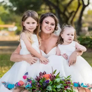 Professional Photographer A smiling bride in a white dress kneels outdoors, hugging two young girls in matching white dresses. Captured by a talented photographer, they pose together with a colorful bouquet, framed by trees and sunlight in the background. www.maranonimagery.com