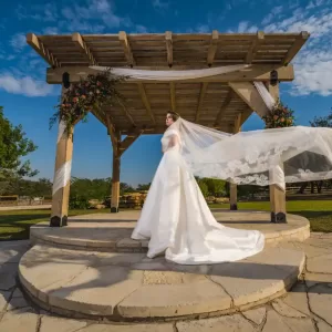 Professional Photographer A bride in a white wedding dress and veil stands on a stone platform under a wooden pergola decorated with flowers, outdoors on a sunny day—perfect for stunning portraits or elegant headshots against the blue sky and scattered clouds. www.maranonimagery.com