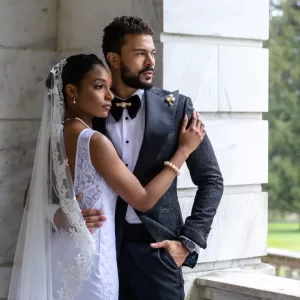 Professional Photographer A bride in a white lace dress and veil embraces a groom in a black tuxedo with gold accents. Captured by a wedding photographer, they stand near a marble wall, gazing thoughtfully into the distance. www.maranonimagery.com