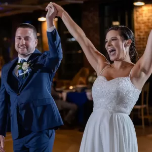 Professional Photographer A smiling bride in a white dress and a groom in a blue suit hold hands and raise their arms in celebration on the dance floor, captured perfectly by the wedding photographer at their lively reception. www.maranonimagery.com