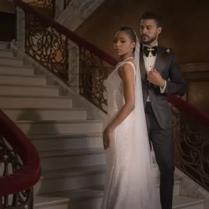 Professional Photographer A bride in a white gown and a groom in a black tuxedo stand together on an elegant staircase with ornate railings, looking confidently at the camera—a timeless moment captured by a skilled wedding photography professional. www.maranonimagery.com
