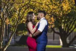Professional Photographer A couple stands close and embraces outdoors, gazing into each other's eyes. Captured by a skilled photographer, the woman in a red dress and man in a blue vest are framed by sunlight and autumn trees for a warm, romantic portrait. www.maranonimagery.com