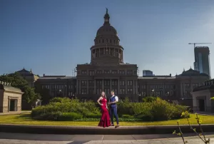 Professional Photographer A woman in a red dress and a man in a blue suit stand together in front of the Texas State Capitol building on a sunny day, captured by a skilled photographer known for stunning headshots. www.maranonimagery.com