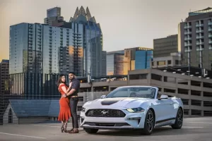 Professional Photographer A couple stands close together in front of a white Ford Mustang convertible on a rooftop parking lot, with modern city skyscrapers and a sunset sky in the background—an unforgettable moment of wedding photography. www.maranonimagery.com