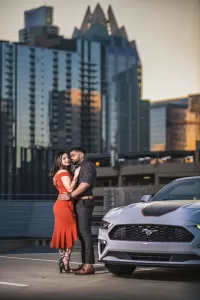 Professional Photographer A couple embraces beside a white Ford Mustang in a city rooftop parking lot, captured by a skilled photographer, with tall glass buildings and a distinctive skyscraper in the background at sunset. www.maranonimagery.com