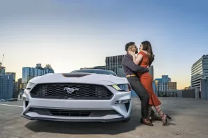 Professional Photographer A couple embraces and smiles while standing next to a white Ford Mustang on a rooftop parking lot with city buildings in the background at sunset, capturing a romantic moment perfect for wedding photography. www.maranonimagery.com