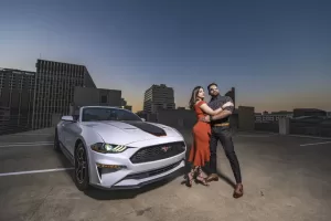 Professional Photographer A stylish couple poses confidently for a photographer on a rooftop parking lot at sunset beside a white Ford Mustang, with city buildings in the background. The woman wears an orange dress and heels; the man wears dark pants and sunglasses. www.maranonimagery.com
