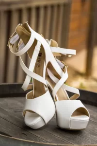 Professional Photographer A pair of white high-heeled, open-toe platform shoes with ankle straps, perfect for wedding photography, displayed on a wooden surface with a blurred background. www.maranonimagery.com