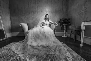 Professional Photographer A bride in a wedding gown and veil sits on a loveseat, smiling with one hand resting on the armrest, in an elegant room with textured walls, wood floor, and a rug—perfect for timeless portraits by a skilled photographer. www.maranonimagery.com