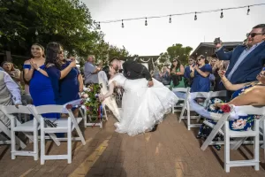 Professional Photographer A groom dips and kisses his bride as they walk down an outdoor aisle lined with cheering guests in formal attire—an unforgettable moment captured by expert wedding photography, with white chairs and string lights overhead. www.maranonimagery.com