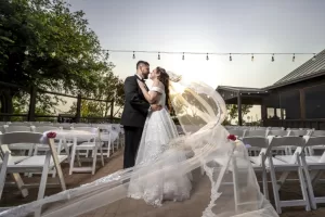 Professional Photographer A bride and groom embrace and kiss in an outdoor wedding venue with empty white chairs. Captured by a skilled wedding photographer, the bride’s long veil flows dramatically in the wind beneath glowing string lights. www.maranonimagery.com