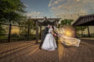 Professional Photographer A bride and groom kiss at sunset under a decorated pergola outdoors. Captured by a skilled wedding photographer, the bride's veil flows in the breeze as they stand on a brick patio surrounded by trees and string lights. www.maranonimagery.com