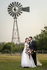 Professional Photographer A bride in a white gown and a groom in a black suit embrace and smile outdoors, holding a bouquet, with a tall windmill and green trees in the background—perfect for romantic wedding photography. www.maranonimagery.com