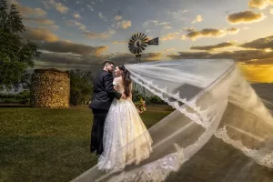 Professional Photographer A bride and groom embrace outdoors at sunset near a stone structure and windmill, with the bride’s long veil flowing dramatically in the wind—a romantic portrait capturing their special moment. www.maranonimagery.com