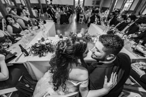 Professional Photographer A bride and groom sit close together, smiling at each other, as a photographer captures portraits of them surrounded by wedding guests seated at long banquet tables in a decorated reception hall. www.maranonimagery.com