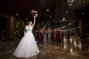 Professional Photographer A bride in a white dress holds a bouquet in the air, ready to toss it to a group of women at an indoor wedding reception with string lights—an unforgettable moment captured through stunning wedding photography. www.maranonimagery.com