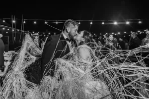 Professional Photographer A photographer captures a bride and groom kissing under string lights at night, surrounded by laughing guests and tangled in long strands of hay or raffia, creating a joyful and festive atmosphere. www.maranonimagery.com