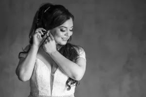 Professional Photographer A smiling bride in a white wedding dress adjusts her earring, looking down. Captured by a skilled photographer, this black and white portrait shows her long, wavy hair and necklace against a softly blurred background. www.maranonimagery.com