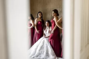 Professional Photographer A bride in a white wedding gown sits surrounded by four bridesmaids in matching red dresses, all posing together indoors for timeless wedding photography, beautifully framed by a blurred doorway. www.maranonimagery.com