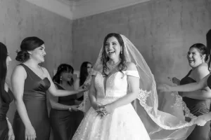 Professional Photographer A bride in a white gown and veil smiles joyfully while bridesmaids in matching dresses laugh and help adjust her veil, as a photographer captures the heartfelt moment in the softly lit room. www.maranonimagery.com