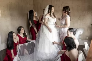 Professional Photographer A bride in a white gown smiles as her bridesmaids, dressed in red, gather around her, holding her veil and laughing together in a warmly lit room—perfect for portraits by an experienced photographer. www.maranonimagery.com