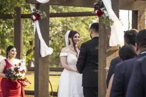 Professional Photographer A bride and groom stand facing each other under a wooden arch decorated with white fabric and flowers during an outdoor wedding ceremony, as guests and a bridesmaid nearby look on—perfect for timeless portraits or candid headshots. www.maranonimagery.com