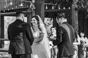 Professional Photographer A bride and groom stand facing each other, smiling, during an outdoor wedding ceremony. The photographer captures their portraits as the officiant stands beside them holding a book, while bridesmaids watch in the background under string lights. www.maranonimagery.com