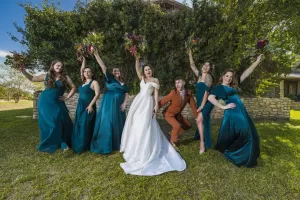 Professional Photographer A bride in a white gown and a groom in a brown suit pose joyfully with five bridesmaids in teal dresses, each holding bouquets and striking playful poses outdoors on a sunny day—captured beautifully by the photographer. www.maranonimagery.com