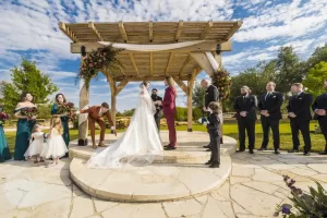 Professional Photographer A wedding ceremony outdoors under a wooden arbor, with the couple holding hands. Bridesmaids and groomsmen stand on either side, children present, and bright blue sky with scattered clouds—perfect for capturing timeless portraits. www.maranonimagery.com