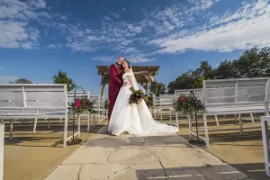 Professional Photographer A bride in a white dress and groom in a maroon suit embrace and smile in an outdoor wedding venue, standing between white benches with flower arrangements under a blue, partly cloudy sky—perfect for romantic wedding photography. www.maranonimagery.com