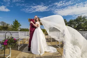 Professional Photographer A bride in a white gown and flowing veil embraces a groom in a burgundy suit outdoors under a bright blue sky, as a photographer captures romantic portraits with empty white benches and floral arrangements behind them. www.maranonimagery.com