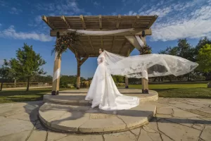 Professional Photographer A bride in a white gown and long veil stands on a stone platform under a wooden pergola decorated with flowers, perfect for stunning wedding photography, with green trees and a blue sky with clouds in the background. www.maranonimagery.com