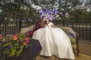 Professional Photographer A newlywed couple sits on a sofa, the person in a maroon suit kisses their partner in a white wedding dress. A neon "This Must Be the Place" sign glows behind them—an intimate moment captured through expert wedding photography. www.maranonimagery.com