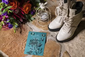 Professional Photographer A teal wedding invitation, two rings, white lace-up boots, a lantern, and a vibrant bouquet with red and purple flowers are artfully arranged on a rustic stone floor—perfect inspiration for creative wedding photography. www.maranonimagery.com
