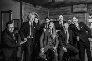 Professional Photographer Eight men in suits pose together indoors, smiling and laughing in a rustic, wooden room. The photographer captures this relaxed, celebratory moment in black and white, creating a timeless group portrait. www.maranonimagery.com